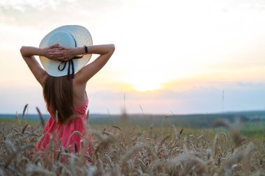 Kırmızı yazlık elbiseli, beyaz hasır şapkalı, sarı tarlada dikilen genç ve mutlu bir kadın. Olgun altın buğdayla kollarını kaldırıp sıcak bir akşamın keyfini çıkarıyor..