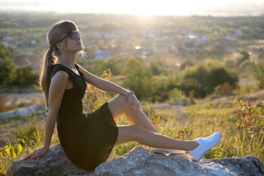 Siyah kısa elbiseli zarif bir kadın yaz akşamı açık havada bir kayanın üzerinde oturmuş dinleniyor. Modaya uygun bir kadın doğada sıcak günbatımının tadını çıkarıyor..