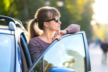 Bir yaz caddesinde arabasının yanında sıcak bir günün tadını çıkaran günlük kıyafetli mutlu bir kadın sürücü. Seyahat ve ulaşım kavramı.