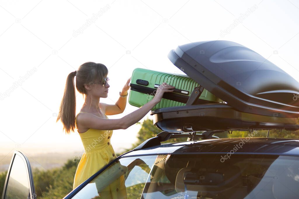 Young woman taking green suitcase out from car roof rack. Travel and vacations concept.