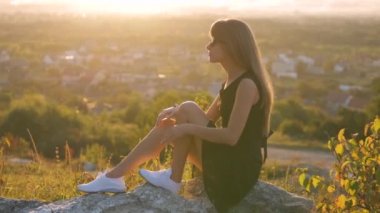 Siyah kısa elbiseli ve beyaz spor ayakkabılı genç zarif bir kadın yaz akşamları bir kayanın üzerinde oturup dinleniyor. Şık bir bayan doğada sıcak günbatımının tadını çıkarıyor..