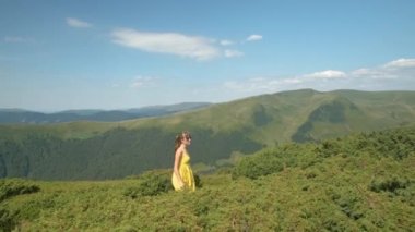 Sarı elbiseli genç bir kadın yaz dağlarında rüzgarlı bir günde çimlerin üzerinde yürüyor..