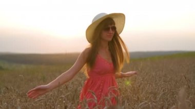 Kırmızı elbiseli mutlu, uzun saçlı genç kadın gün batımında altın buğday tarlasında dönüyor..