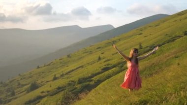 Kırmızı elbiseli mutlu bir kadın yürüyüşçünün arka görüntüsü çimenli bir tepede rüzgarlı bir sonbahar akşamında kollarını açıp güneşin batışının keyfini çıkarırken..