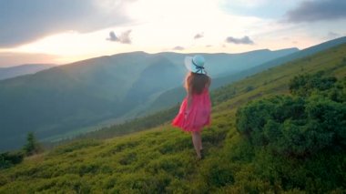 Kırmızılı genç kadın yaz dağlarında rüzgarlı bir akşamda yamaçtaki çayırda yürüyor..