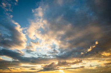 Turuncu batan güneş ve mavi gökyüzü tarafından aydınlatılan kabarık bulutlarla dramatik günbatımı manzarası.