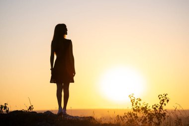 Yazlık elbiseli, karanlık bir kadın silueti açık havada duruyor gün batımında manzaranın tadını çıkarıyor..