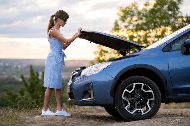 Kaputu açık, bozuk bir arabanın yanında duran genç bir kadın sürücü aracın motorunu kontrol ediyor ve yardım bekliyor..