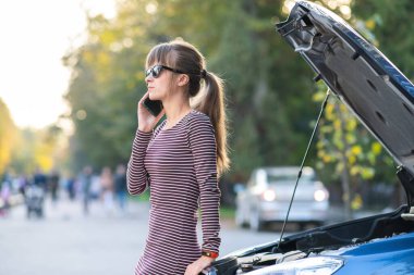 Sinirli genç kadın sürücü cep telefonuyla konuşuyor. Şehir caddesinde kaputu açık bozuk bir arabanın yanında yardım servisi var..