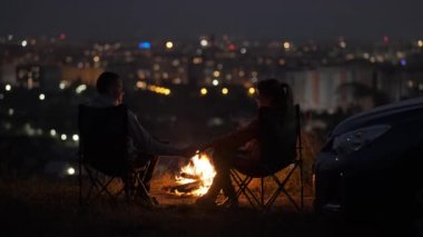 Geceleri yanan sarı kamp ateşinin yanında kamp sandalyelerinde oturan mutlu genç çift..