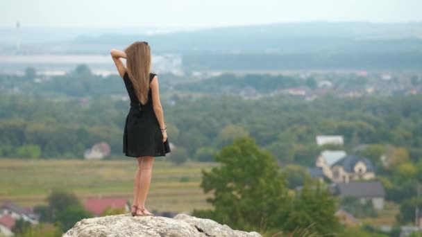 丘の上に立つ短い黒のドレスで長い髪の若い女性は暖かい夏の日を楽しむ — ストック動画