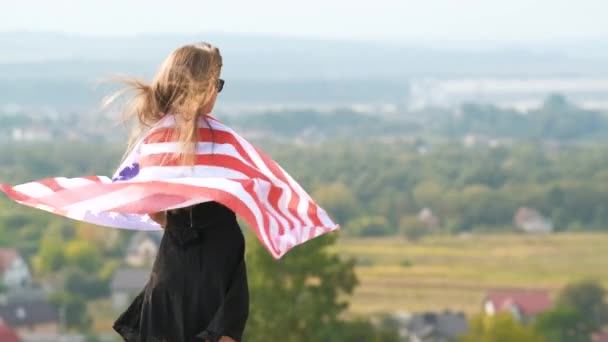 夏の暖かい日を楽しむ屋外でリラックス彼女の肩の上に風アメリカ国旗に手を振って長い髪を保持している若い幸せなアメリカ人女性 — ストック動画