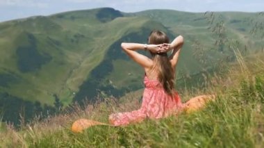 Kırmızı elbiseli genç kadın yeşil çimenlikte dinleniyor ılık güneşli bir günde yaz dağlarında doğa manzarasının tadını çıkarıyor..