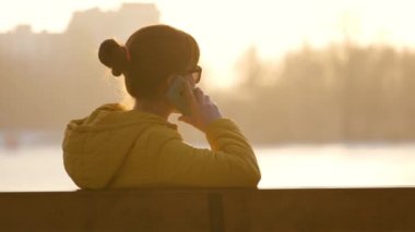 Parktaki bankta oturan genç ve güzel bir kadın sıcak bir sonbahar akşamında dışarıda akıllı telefonuyla konuşuyor..