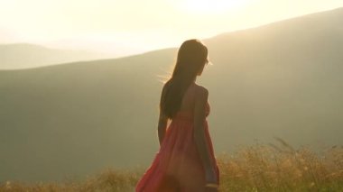 Kırmızılı genç kadın yaz dağlarında rüzgarlı bir akşamda yamaçtaki çayırda yürüyor..