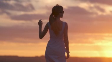 Yazlık elbiseli, karanlık bir kadın silueti açık havada duruyor gün batımında manzaranın tadını çıkarıyor..