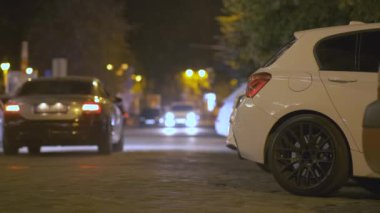 Gece yol kenarındaki park halindeki arabaları kapatın. Şehir caddesinde hareket halindeki araçların trafik ışıklarının bulanık görüntüsüyle..