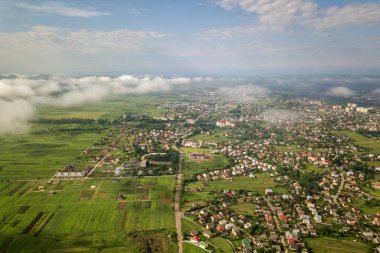 Yazın yeşil tarlalar arasında sıralı binalar ve kıvrımlı sokaklar olan bir kasaba ya da köyün üstündeki beyaz bulutların hava görüntüsü. Yukarıdan kırsal alan.