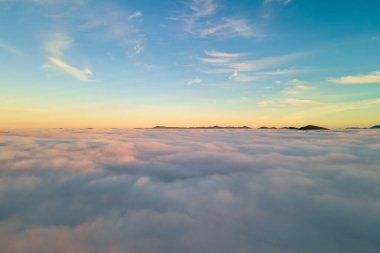 Ufuktaki uzak karanlık dağlarla beyaz yoğun bulutların üzerinde canlı günbatımının havadan görünüşü.