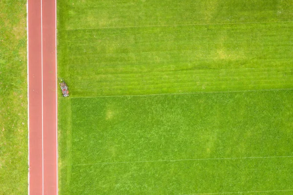 緑の草を刈る労働者の小像の空中図サッカー競技場フィールド上の芝刈り機と夏に赤い走路 — ストック写真