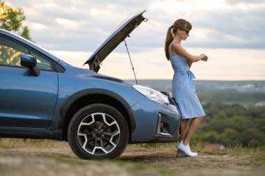Kaputu açık, bozuk bir arabanın yanında duran genç bir kadın sürücü aracın motorunu kontrol ediyor ve yardım bekliyor..