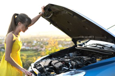 Arabasının yanında duran kafası karışmış kadın sürücü kaputu patlamış, bozuk motoru inceliyor..