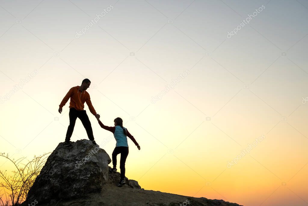Man and woman hikers helping each other to climb a big stone at sunset in mountains. Couple climbing on a high rock in evening nature. Tourism, traveling and healthy lifestyle concept.