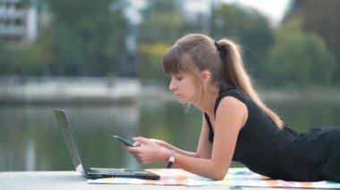 Genç bir kadın cep telefonuyla konuşurken dizüstü bilgisayarın arkasında çalışıyor yaz parkında yatıyor..