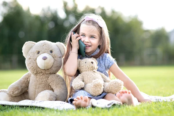 一个漂亮的小女孩和她的玩具熊一起坐在公园里 一边用手机聊天 一边在户外快乐地微笑着 — 图库照片