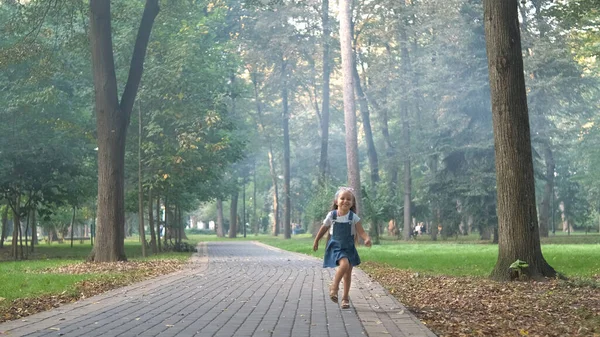 穿着粗斜纹棉布衣服的快乐微笑的小女孩在夏日公园的人行道上奔跑 — 图库照片