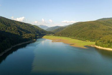 Yoğun yeşil ormanlarla kaplı yüksek dağ tepeleri arasında berrak mavi suyu olan büyük göl manzarası..