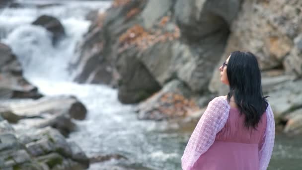 一位黑发女子身穿长裙 穿着时髦的晚礼服 坐在小山小河边的石头上 流水湍急流淌 — 图库视频影像