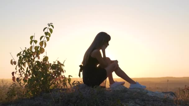 夏天的傍晚 穿着黑色短裙和白色运动鞋的年轻优雅女人坐在岩石上放松自己 时尚的女士享受大自然温暖的落日 — 图库视频影像