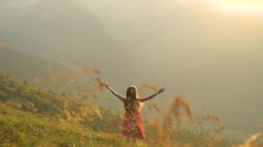 Kırmızı elbiseli genç mutlu kadın yolcunun arka görüntüsü. Çimenli bir yamaçta duruyor. Yazın rüzgarlı bir akşamda, kollarını açıp güneşin batışının keyfini çıkarıyorlar..