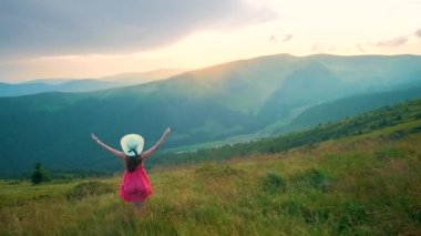Kırmızı elbiseli genç mutlu kadın yolcunun arka görüntüsü. Çimenli bir yamaçta duruyor. Yazın rüzgarlı bir akşamda, kollarını açıp güneşin batışının keyfini çıkarıyorlar..