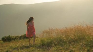Kırmızılı genç kadın yaz dağlarında rüzgarlı bir akşamda yamaçtaki çayırda yürüyor..