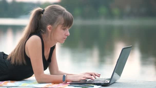屋外の夏の公園に寝そべっている携帯電話に話をしながら ラップトップコンピュータの後ろで働く若い女性オフィスの従業員 リモートワークの概念 — ストック動画