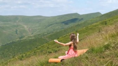 Kırmızılı genç kadın çimenli bir tarlada oturuyor rüzgarlı bir yaz gününde, dağlarda doğa manzarasının tadını çıkarıyor..
