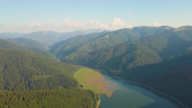 Yoğun yeşil ormanlarla kaplı yüksek dağ tepeleri arasında berrak mavi suyu olan büyük göl manzarası..