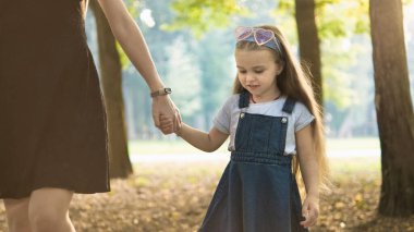 Genç anne ve uzun saçlı küçük kızı yaz parkında el ele yürüyorlar..