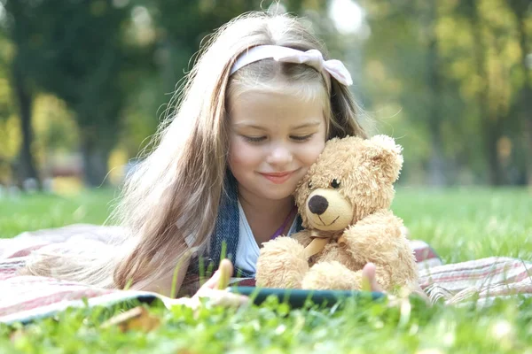 快乐的小女孩在夏天的公园里和最喜欢的玩具熊一起玩她的手机 — 图库照片