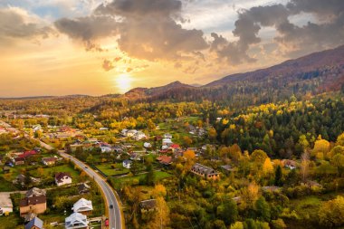 Günbatımında sarı ve yeşil çam ağaçlarıyla kaplı sonbahar dağları arasında küçük evleri olan kırsal bir köyün havadan görünüşü..