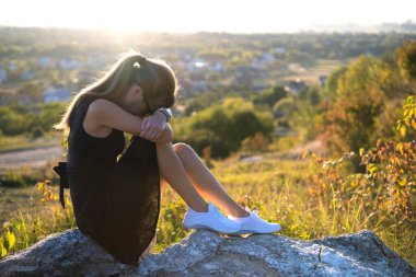 Siyah kısa yaz elbisesi içinde depresif genç bir kadın gün batımında bir kayanın üzerinde oturup açık havada düşünüyor. Modaya uygun bir kadın doğada sıcak bir akşamda tefekkür ediyor..