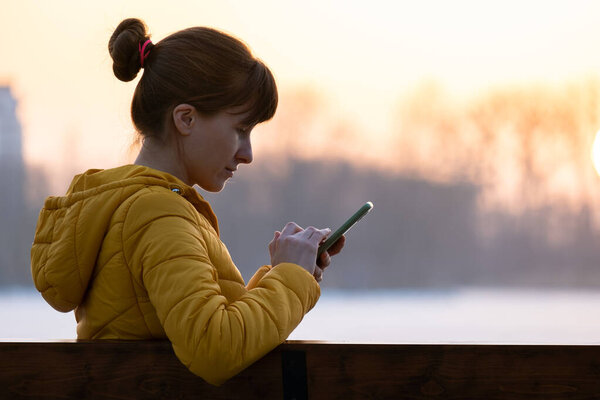 Young pretty woman sitting on a bench browsing her mobile phone outdoors in the evening.