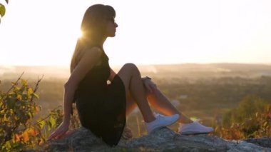 Siyah kısa elbiseli ve beyaz spor ayakkabılı genç zarif bir kadın yaz akşamları bir kayanın üzerinde oturup dinleniyor. Şık bir bayan doğada sıcak günbatımının tadını çıkarıyor..