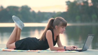 Laptopun arkasında çalışan genç bir kadın dışarıda, yaz parkında yatıyor..