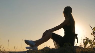 Siyah kısa yaz elbisesi içinde genç ve güzel bir kadın gün batımında bir kayanın üzerinde oturup dinleniyor. Modaya uygun bir kadın doğada sıcak bir akşamın tadını çıkarıyor..