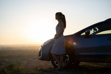 Mavi elbiseli mutlu genç kadın sürücü sıcak yaz akşamının tadını çıkarıyor arabasının yanında duruyor. Seyahat ve tatil kavramı.