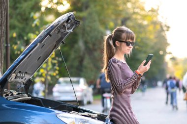 Kaputu açık, bozuk bir arabanın yanında cep telefonuyla konuşan genç üzgün bir kadın sürücü şehir caddesinde aracıyla sorun yaşaması için yardım bekliyor..