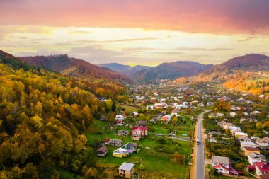 Günbatımında sarı ve yeşil ladin ormanlarıyla kaplı sonbahar dağları arasında küçük evleri olan köyün kırsal manzarası..